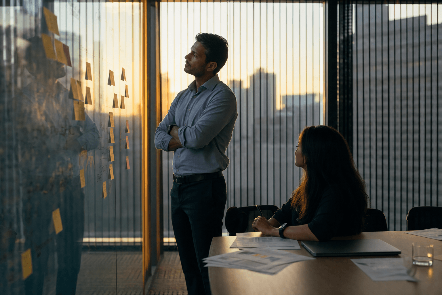Director de marketing con los brazos cruzados frente a una pared de cristal cubierta de post-its, junto a un colega con una hoja de cálculo impresa en una oficina al anochecer