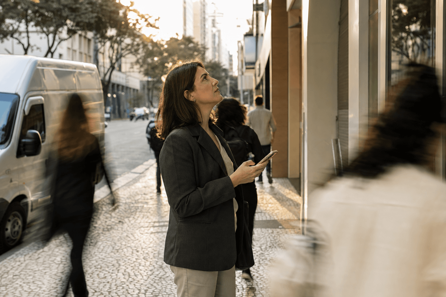 Mujer con blazer en la acera de una ciudad mirando el móvil mientras observa la entrada de un edificio comercial al final de la tarde