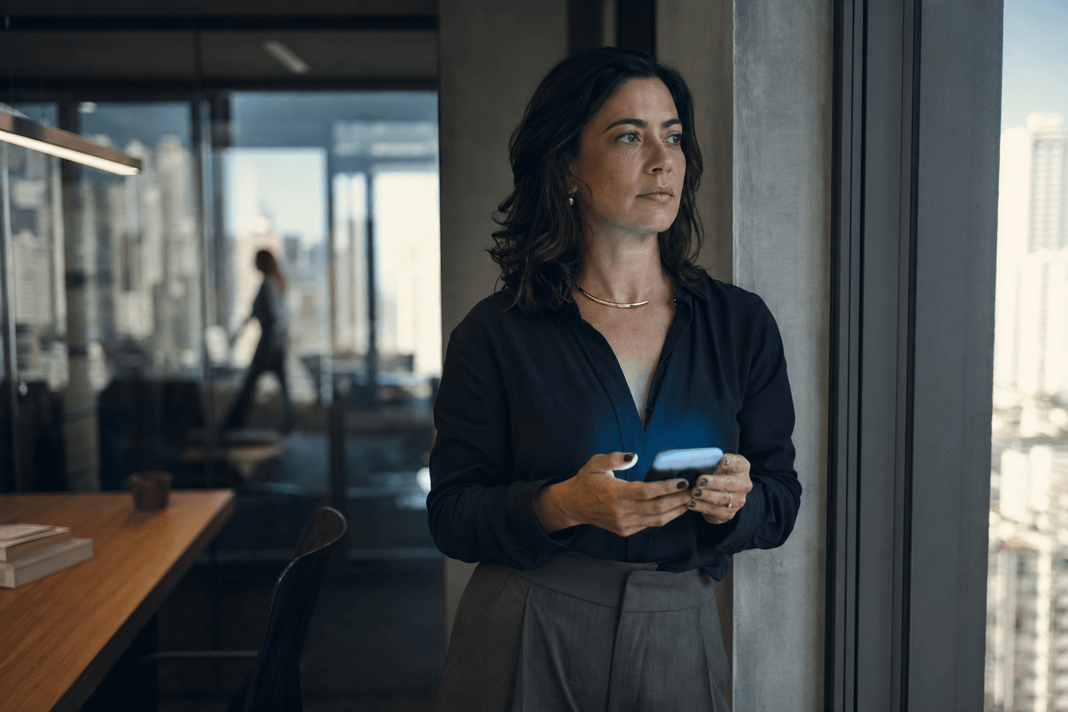 Directora de marketing frente a la ventana de una oficina en São Paulo, sosteniendo el móvil cuya pantalla ilumina su rostro.