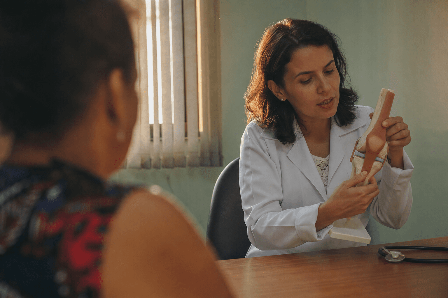 Médica con bata blanca sostiene un modelo anatómico de rodilla mientras conversa con un paciente en una consulta iluminada con luz natural